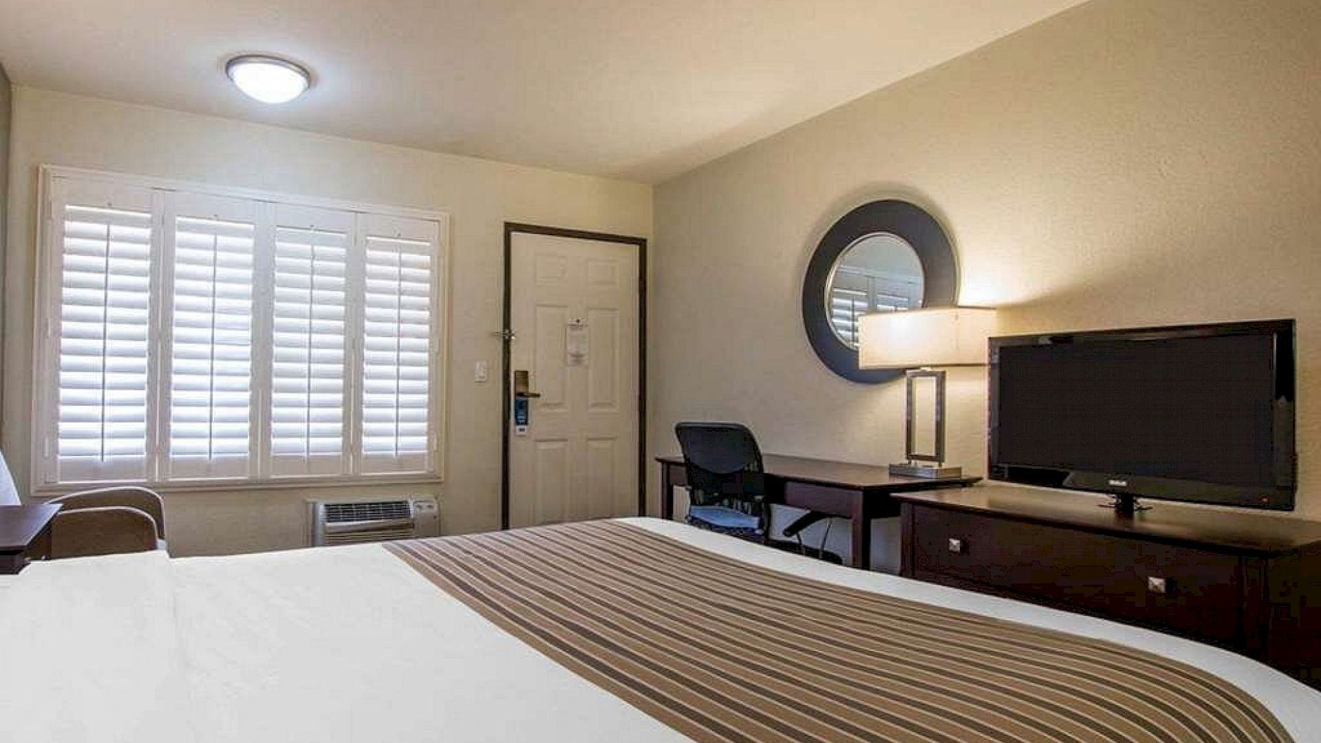 A bright office-style bedroom with a bed, shuttered window, desk and monitor, wall mirror, and overhead light.